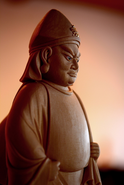 仏像彫刻原田謹刻 大黒天像(だいこくてんぞう) 【大黒天】 元来はヒンズー教の神で、密教では大自在天の化身である。サンスクリット語のマハーカーラの訳で、摩訶迦羅と音写する。マハーカーラは偉大な黒い神、偉大な時間=破壊者を意味する。マハーとは大もしくは偉大なる、カーラとは時あるいは黒を意味する。三宝を守護し、戦闘をつかさどり、飲食を豊かにする神で黒色忿怒相を示す。中国南部では金袋を持ち腰掛ける姿となり、諸寺の厨房に祀られた。わが国の大黒天はこの系統で、最澄によってもたらされ、天台宗の寺院を中心に祀られたのがその始まりといわれる。その後、台所の守護神から福の神としての色彩を強め、七福神の一つとなり、頭巾をかぶり左肩に大袋を背負い、右手に小槌を持って米俵を踏まえるといった現在よくみられる姿になる。音韻や容姿の類似から大国主命と重ねて受け入れられ、商売繁盛を願う商家はもとより、農家においても田の神として信仰を集めている。