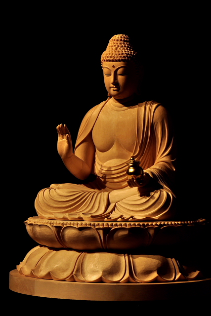 仏像彫刻原田謹刻 薬師瑠璃光如来坐像(やくしるりこうにょらいざぞう) 【薬師瑠璃光如来】 東方浄瑠璃世界の教主。 人々を病の苦しみから救い、寿命を延ばし、貧困を除く。 薬師様は、まだ菩薩として修行しておられた時に衆生を救済する十二の大願を立てられ、それを成就されて如来になられました。薬師様には、病からの救済のほか、古くは罪を懺悔(さんげ)する修法の本尊として、また、天変地異や政変などの社会不安からの救済という大きな願いが託されてきました。左手には万病に効く薬が入った薬壺(やっこ)を持たれています。「薬師」とは、外科が存在しなかった中世以前には、現代の医師に対応する言葉であったそうです。