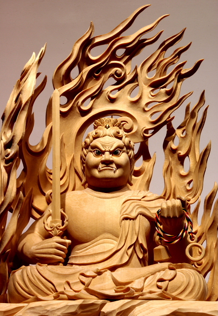 仏像彫刻原田謹刻 不動明王坐像(ふどうみょうおうざぞう) 【不動明王】 悪を降(くだ)し、衆生(しゅじょう)を守る。仏道に導きがたいものを畏怖せしめ、煩悩を打ちくだく。菩提心の揺るがないことから不動という。大日如来の化身として、すべての悪と煩悩をおさえしずめ、生あるものをすくう。岩上に座して火炎に包まれた姿で、怒りの形相に表す。右手に悪をたちきる剣を、左手に救済の索を持つ。サンスクリット名アチャラナータAcalanāthaの漢訳で、発音に従い阿遮羅囊他と記す場合もある。 ◆このお姿は、一般在家のお客様からのご注文で納入させていただいたお姿です。御面相や体躯、光背の形状などは、お客様がお持ちの複数のお写真を基に、ご希望を伺った上で謹刻させていただきました。