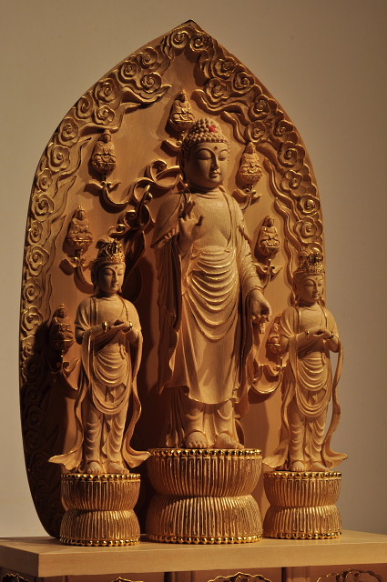 仏像彫刻原田謹刻 一光三尊阿弥陀如来像 【善光寺式阿弥陀三尊】 阿弥陀如来を中尊に,その左右に観音、勢至の二菩薩を脇侍とする。三尊全体の背後を大きな一枚の舟形光背がおおっている。これにより「一光三尊」という。この一光三尊形式の源流は中国北魏時代の仏像に求められる。三尊足下の蓮台は臼形蓮台とよばれ,蓮肉部の高い垂敷蓮華である。光背は大きな舟形光背で,光中に七化仏がある。 ◆お施主様からのご要望で、総高45㎝のお姿を謹刻納入させていただきました。桧の白木素地に、金線が施してございます。